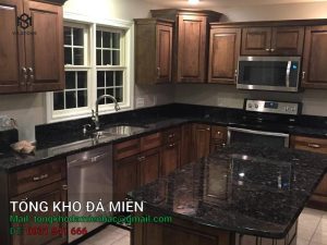 Đá bàn bếp Granite Volga Blue còn được biết đến với tên gọi Xà Cừ Ngọc Trai, là một trong những loại đá granite cao cấp được ưa chuộng trên toàn thế giới. Đá được khai thác chủ yếu tại vùng Golovinske, khu vực Zhitomir, Ukraine. Với màu sắc độc đáo và vẻ đẹp tự nhiên, Volga Blue mang đến vẻ đẹp sang trọng và đẳng cấp cho nhiều hạng mục trang trí nội thất và ngoại thất. Xuất xứ của đá bàn bếp granite Volga Blue Đá Volga Blue được khai thác từ nhiều mỏ khác nhau xung quanh khu vực Golovinske, Zhitomir, Ukraine. Mỗi mỏ đá mang lại những tấm đá với màu sắc và chi tiết vân không hoàn toàn giống nhau, tạo nên sự đa dạng và độc đáo cho từng tấm đá. Đặc điểm của đá bàn bếp granite Volga Blue Volga Blue nổi bật với sự pha trộn giữa màu đen, xanh đậm và xám. Đặc biệt, khi được chiếu sáng, đá ánh lên những hạt khoáng sản lấp lánh, tạo nên hiệu ứng xà cừ ngọc trai vô cùng bắt mắt. Đây là loại đá có hạt khoáng sản to, là một loại khoáng chất anorthosite màu đen, xanh đậm, xám và ánh lên khi gặp ánh sáng. Thành phần khoáng học Đá Volga Blue là một loại anorthosite, chứa nhiều felspat plagioclase (90-100%) cùng với các khoáng chất mafic như pyroxen, ilmenit, magnetit, và olivin. Thành phần khoáng học của đá bao gồm: Plagioclase: 85% Quartz: 7% Pyroxene: 3% Orthoclase: 3% Biotite: 1% Accessories: 1% Thông số kỹ thuật Khả năng hấp thụ nước: 0.07 - 0.09% khối lượng đá. Cường độ nén: 190.70 MPa Khối lượng riêng: 2623 kg/m³ Độ dày: 20mm Bề mặt hoàn thiện Đá Granite Volga Blue có thể được hoàn thiện với các bề mặt sau: Polished (Bóng): Bề mặt đá được đánh bóng, tạo nên độ sáng bóng và phản chiếu ánh sáng đẹp mắt. Honed (Mờ): Bề mặt đá mờ, không bóng, mang lại vẻ đẹp tự nhiên và mộc mạc. Leather (Da): Bề mặt đá có độ nhám nhẹ, tạo cảm giác mềm mại và độc đáo khi chạm vào. Ứng dụng của Đá Bàn Bếp Granite Volga Blue Nhờ vẻ đẹp sang trọng cùng những tính năng ưu việt, đá bàn bếp granite Volga Blue có thể ứng dụng cho cả không gian nội thất và ngoại thất. Nếu biết cách kết hợp với đồ nội và ngoại thất, bạn có thể tạo nên một không gian sống tuyệt đẹp. Ốp bếp Đá Volga Blue là sự lựa chọn hoàn hảo cho bề mặt bếp nhờ màu sắc độc đáo và độ bền cao. Với sự pha trộn của các tông màu đen, xanh đậm và xám, đá tạo ra một không gian bếp hiện đại và tinh tế. Không chỉ mang lại vẻ đẹp sang trọng, bề mặt đá còn dễ dàng vệ sinh, chống thấm nước và chịu được nhiệt độ cao, giúp cho công việc bếp núc trở nên thuận tiện và an toàn hơn. Ốp cầu thang Ứng dụng đá Volga Blue cho cầu thang giúp tạo nên vẻ đẹp mạnh mẽ và bền bỉ. Đặc tính chống mài mòn và khả năng chịu lực tốt của đá granite đảm bảo cho cầu thang có độ bền cao và mang lại cảm giác an toàn cho người sử dụng. Màu sắc và vân đá tinh tế cũng làm tăng thêm sự sang trọng cho không gian sống. Ốp mặt tiền Volga Blue là lựa chọn xuất sắc để ốp mặt tiền nhà, tạo nên điểm nhấn ấn tượng và độc đáo. Màu sắc đá kết hợp với ánh sáng tự nhiên tạo ra hiệu ứng thị giác tuyệt đẹp, thu hút ánh nhìn từ mọi góc độ. Bề mặt đá bền vững với thời gian, giúp mặt tiền ngôi nhà luôn giữ được vẻ đẹp nguyên bản, bất chấp các tác động của thời tiết và môi trường. Ốp tường ngoại thất Với khả năng chống chịu thời tiết và độ bền vượt trội, đá bàn bếp granite Volga Blue cũng được ứng dụng rộng rãi trong các hạng mục ngoại thất. Đá còn giữ được màu sắc và độ bóng lâu dài. Vì vậy, đây là lựa chọn lý tưởng để tạo ra một bức tường ngoại thất đẹp mắt và bền vững, giúp nâng cao giá trị thẩm mỹ và bảo vệ công trình khỏi các tác động xấu từ môi trường. Nếu có điều kiện, bạn có thể sử dụng đá Volga Blue để lát sân vườn mang lại vẻ đẹp tự nhiên và sang trọng cho không gian ngoài trời. Đá granite với độ bền cao, khả năng chống trượt và dễ dàng vệ sinh, giúp sân vườn luôn sạch sẽ và an toàn cho người sử dụng. Màu sắc và hoa văn đặc biệt của đá Volga Blue tạo nên một không gian sân vườn hài hòa với thiên nhiên, mang lại cảm giác thư thái và dễ chịu. Đá Granite Volga Blue (Xà Cừ Ngọc Trai) là lựa chọn hoàn hảo cho cả nội thất và ngoại thất nhờ vào vẻ đẹp độc đáo, độ bền cao và tính năng ưu việt. Từ bề mặt bếp, cầu thang, mặt tiền nhà đến tường ngoại thất và sân vườn, Volga Blue mang lại sự tiện lợi và bền vững cho các công trình kiến trúc. Lưu ý khi sử dụng Do đặc tính dễ vỡ của đá Volga Blue, khi sử dụng chúng ta cần phải gia cố lại bằng tấm nhựa hoặc sợi thủy tinh để đảm bảo độ bền và an toàn của đá. Đặc biệt, khi sử dụng cho các dự án lớn, cần đảm bảo các tấm đá đều được khai thác từ cùng một mỏ để đảm bảo tính đồng nhất về màu sắc và đường vân. Đá Granite Volga Blue (Xà Cừ Ngọc Trai) là lựa chọn lý tưởng cho những ai yêu thích vẻ đẹp tự nhiên và sự sang trọng. Với màu sắc độc đáo, khả năng chống chịu tốt và đa dạng ứng dụng, Volga Blue chắc chắn sẽ mang lại sự hài lòng cho mọi khách hàng. Để tìm hiểu thêm về đá bàn bếp granite Volga Blue, hãy liên hệ với Tổng Kho Đá Miền Bắc. Chúng tôi sẽ tư vấn chi tiết và báo giá tốt nhất cho sản phẩm đá Granite Volga Blue.